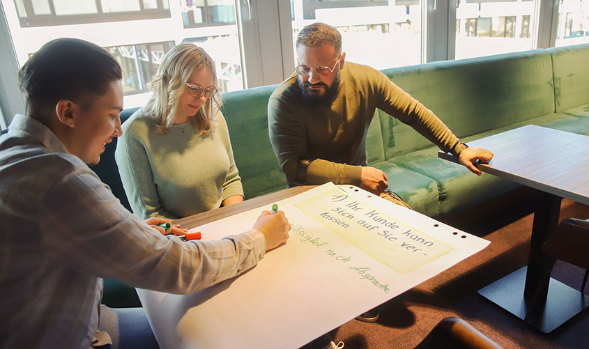 Drei Personen am Tisch gebeugt über ein großen Blatt Papier vom Flipchart
